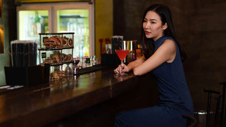 Nightlife concept a pretty girl with long hair wearing blue dress holding a pink drink appreciating the musics alone in the bar.の写真素材