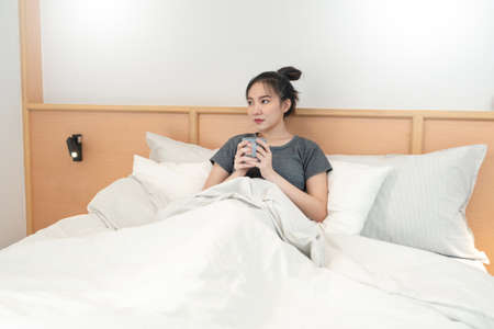 Bedroom concept in the bed a girl appreciating a scent and a taste of the hot coffee in her hands gazing outside the window.の写真素材
