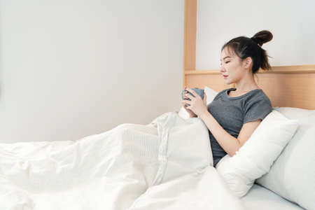 Bedroom concept in the bed a girl appreciating a scent and a taste of the hot coffee in her hands gazing outside the window.の写真素材