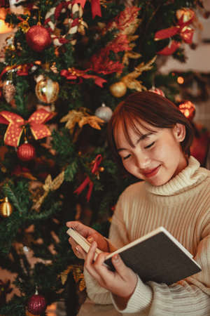 a young Woman in front of Christmas tree reading a book at home with x-mas decorationの写真素材