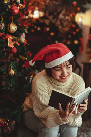 a young Woman in front of Christmas tree reading a book at home with x-mas decorationの写真素材