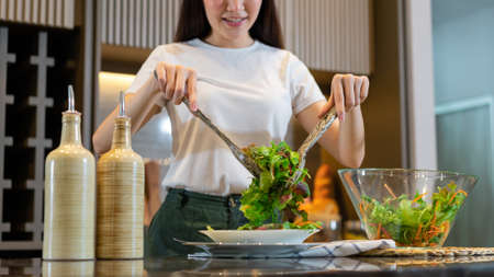 salad concept the woman with white t-shirt and dark green pants standing at the kitchen and making a bowl of green salad.の写真素材