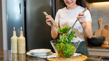 salad concept the woman with white t-shirt and dark green pants standing at the kitchen and making a bowl of green salad.の写真素材
