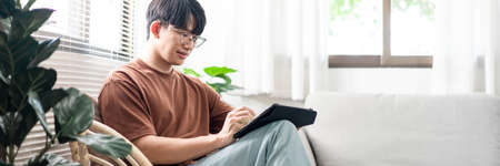 Technology Concept The young man who is sitting on the chair holding his tablet and noting something on it.の写真素材