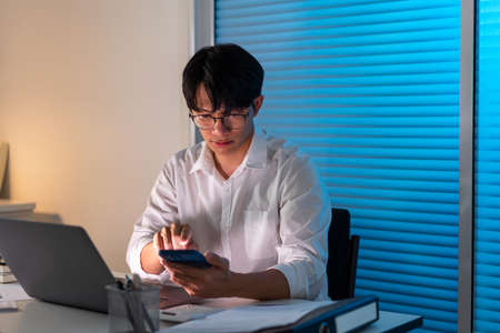 Overwork Concept The office man who sits at his desk holding his smartphone for searching some information.の写真素材