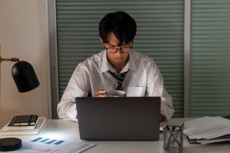 Overwork Concept An office man who works late drinking a cup of coffee while concentrating on his work on the notebook.の写真素材