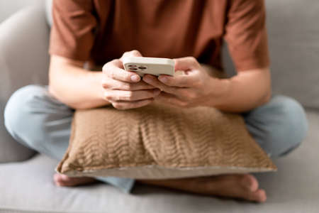 Technology Concept The man sitting next to a pot of plant, putting a small pillow on his lap, and checking something on his smartphone.の写真素材