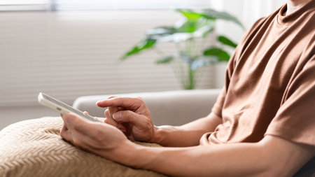 Technology Concept. The man sitting next to a pot of plant, putting a small pillow on his lap, and checking something on his smartphone.の写真素材