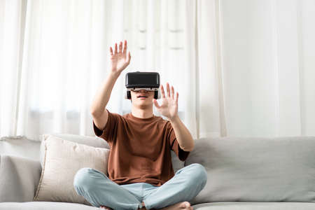 Technology Concept The man with his brown T-shirt and light blue jeans wearing a virtual reality headset while sitting on the sofa.の写真素材