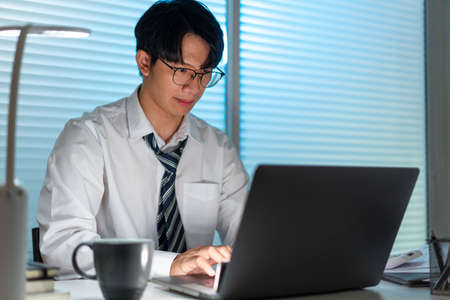 Overwork Concept The office personnel in the loose tie sitting at his desk, reading a paperwork and typing on the notebook.の写真素材