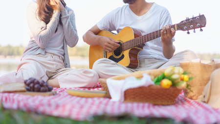 Young man playing acoustic guitar for girlfriend while sitting on blanket to picnic time together.の写真素材