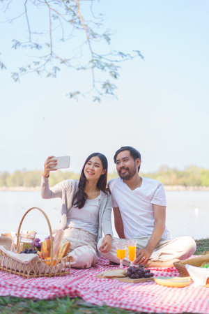 Young couple in love sitting on blanket with food for picnic time and selfie on smartphone together.の写真素材