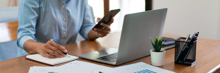 Close up of businesswoman is searching data on smartphone while working on laptop and writing notes.