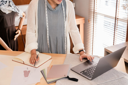 Asian tailor women checking data on laptop to design fashion collection and sketching in notebook.
