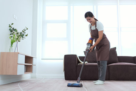 Maid holds wireless cordless vacuum cleaner to vacuuming dust and cleaning on carpet in living room.