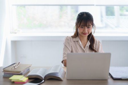 Young asian student in casual clothes wearing earphones to watching tutorial and studying lecturer education online class on laptop while writing knowledge data and working homework in university.の写真素材