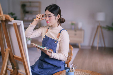 Young asian female artist touching glasses and looking on canvas to thinking idea painting artwork.の写真素材