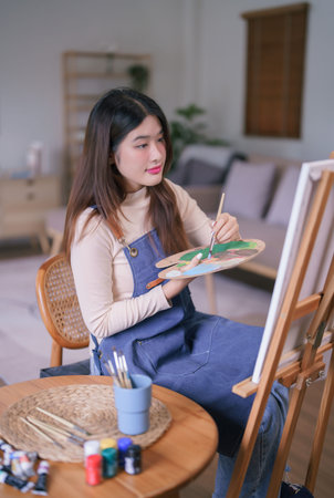 Young asian female artist using paintbrush to mixing color on palette to create painting on canvas.の写真素材