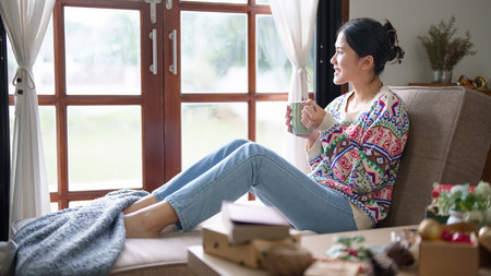 Young asian woman in sweater looking outside the window with dreamy while sitting on the comfort couch and drinking hot chocolate with marshmallow in living room decorating christmas ornaments.の写真素材