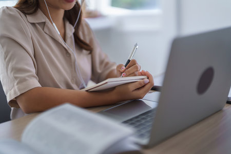 Young asian student in casual clothes writing knowledge data in notebook while wearing earphones to watching tutorial and studying lecturer education online class with laptop technology in university.の写真素材