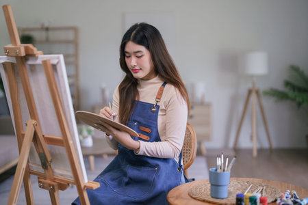 Young asian female artist using paintbrush with color palette to create painting artwork on canvas.の写真素材
