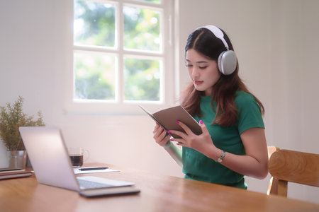 Young asian student women wearing headphone to studying and watching lecture webinar in online class on laptop while reading a book and learning knowledge remote education from home in living room.の写真素材