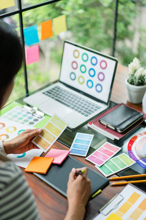 Creative graphic designer comparing color swatch on paper and laptop to designing project on tablet.