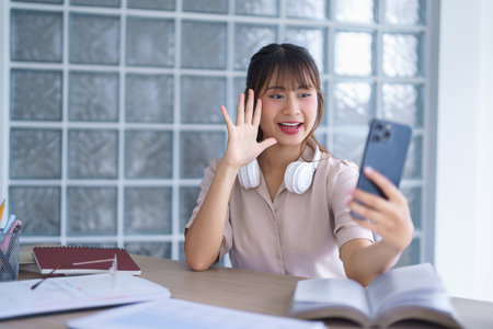 Young asian student in casual clothes wear headphone in her neck while greeting and talking with friends in video call on smartphone after studying education knowledge to doing homework in university.の写真素材