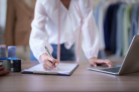 Young asian merchant women wearing measure tape in her neck and checking purchase online order on laptop to taking notes in clipboard while working about online shopping business in home office.の写真素材