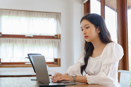 Young asian businesswoman typing business report on laptop and reading financial data on tablet while analysis strategy about investment and planning of new startup project to working in cafe.の写真素材