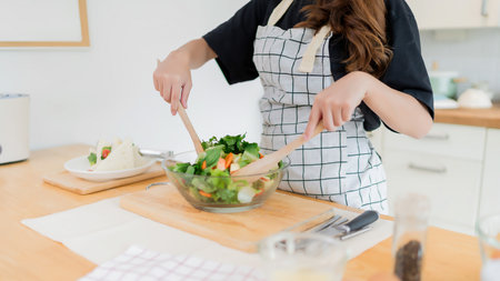 Young asian woman mixing fresh salad vegetables in glass bowl with spoon and cooking delicious sandwich for healthy breakfast food and homemade snack in modern kitchen with healthy lifestyle at home.の写真素材