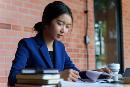 A businesswoman in a blue suit working at a desk with a laptop, holding a coffee cup. She is focused...