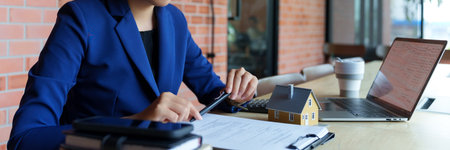 A businesswoman in a blue suit is sitting at a desk, focused on reviewing documents. The background features a brick wall. On the deskの写真素材