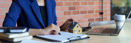 A businesswoman in a blue suit working at a desk with a laptop, holding a pen and writing on a document. A small model house and coffee cup are placed on the desk.の写真素材