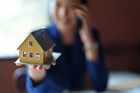 A house seller in a blue suit is holding a model house while talking on a phone. She is smiling and seems engaged in a conversation.の写真素材