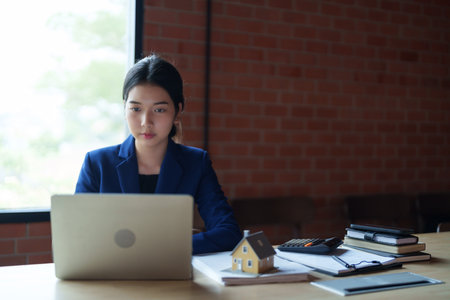 A house seller in a blue suit is working on her laptop at a desk. The background features a brick wall. On the desk, there is a small model house along with some booksの写真素材
