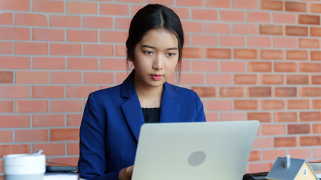 A house seller in a blue suit is working on her laptop at a desk. The background features a brick wall. On the desk, there is a small model house along with some booksの写真素材