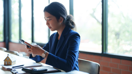 A house seller in a blue suit is focused on her phone while sitting at a desk. The bright, natural...