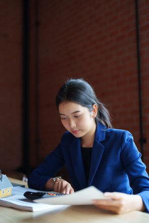 A house seller in a blue suit is working with a calculator at a desk. She is holding a piece of...