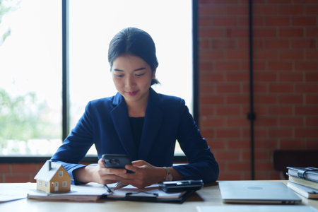 A house seller in a blue suit is focused on her phone while sitting at a desk. The bright, natural...