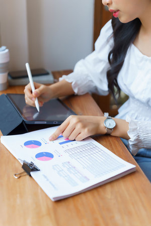 Closeup hands of young asian businesswoman checking finance data on business document and taking notes on tablet while thinking about investment and planning of new startup project to working in cafe.の写真素材