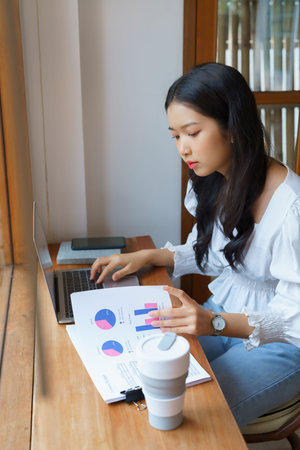 Young asian businesswoman checking marketing data of business on document and typing data on...