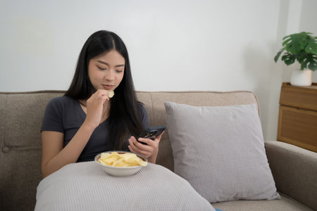 Young asian women using smartphone to surfing social media entertainment and eating snack with...