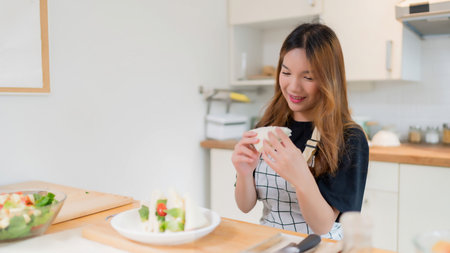 Young asian woman having breakfast and eating delicious sandwich after making to cooking fresh vegetables salad and homemade snack for healthy food in modern kitchen with healthy lifestyle in home.の写真素材