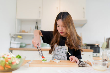 Young asian woman using tongs to cooking delicious sandwich and fresh vegetables salad for healthy breakfast food to preparing homemade snack cutting board in modern kitchen with healthy lifestyle at home.の写真素材