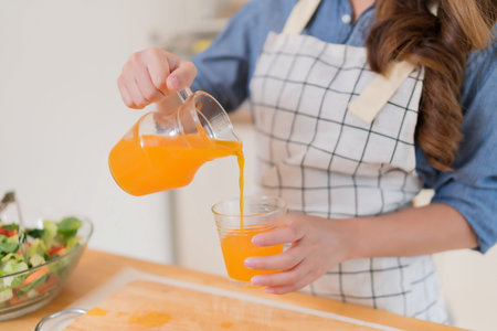 Closeup hands of young asian woman pouring orange juice from jug into the glass while preparing vegetables salad and cooking healthy breakfast food in modern kitchen with healthy lifestyle at home.の写真素材