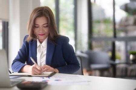 Asian businesswomen reading marketing business report to checking financial chart on laptop and taking notes in notebook while analyze investment and market stock of new startup in outside office.の写真素材