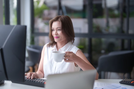 Asian businesswomen drinking coffee and typing information on keyboard to working about planning of marketing report on computer while analyze investment and market stock of new startup in workplace.の写真素材