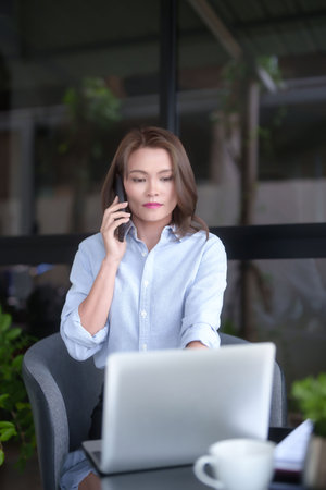 Asian businesswomen taking with customer on smartphone and typing information on laptop to working about planning of marketing business report while analyze investment and market stock of new startup.の写真素材