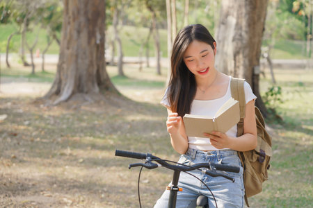 Young asian women with backpack sitting on bicycle and reading a book with happiness to resting after cycling bicycle in the nature park while relaxation with journey travel for healthy lifestyle.の写真素材